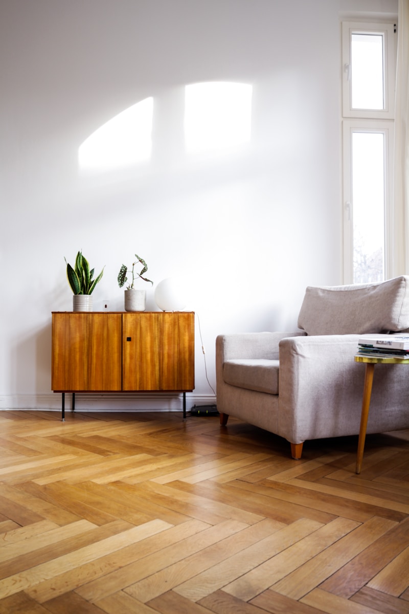 Bright, minimalist living room featuring a wooden sideboard with plants and a soft gray armchair beside a geometric wooden floor.